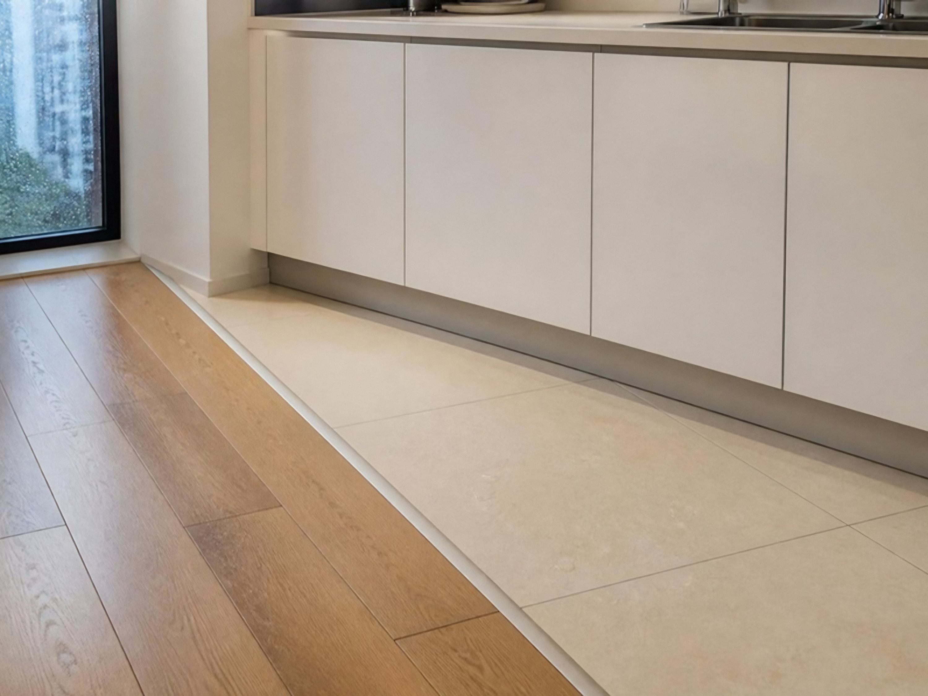 AI image of an oak-look vinyl floor meeting cream kitchen tiles at a neat metal strip below white cabinets